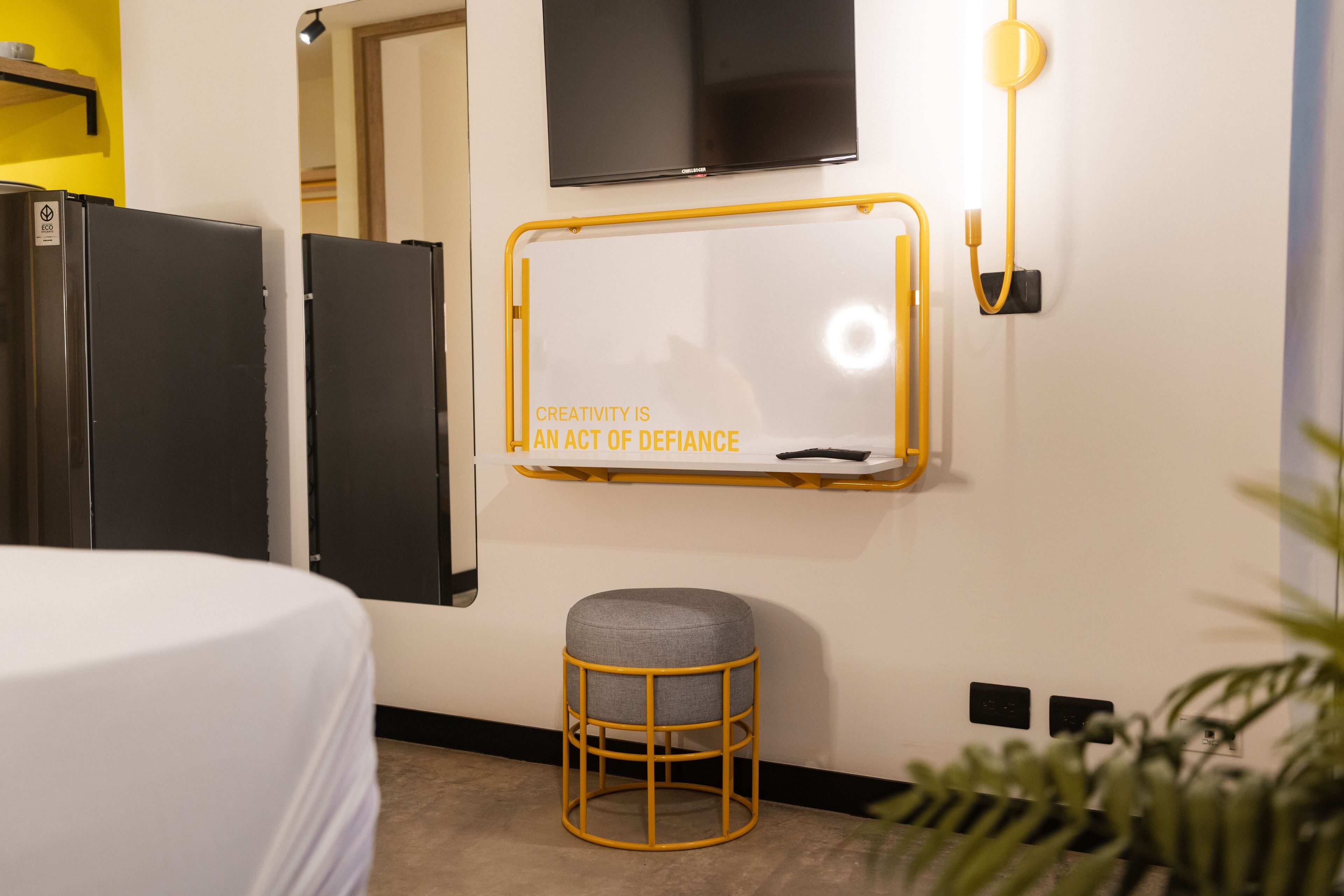 Habitación de hotel con cama, una pizarra con cita motivacional, dos refrigeradores negros, un taburete pequeño y un televisor montado en la pared.