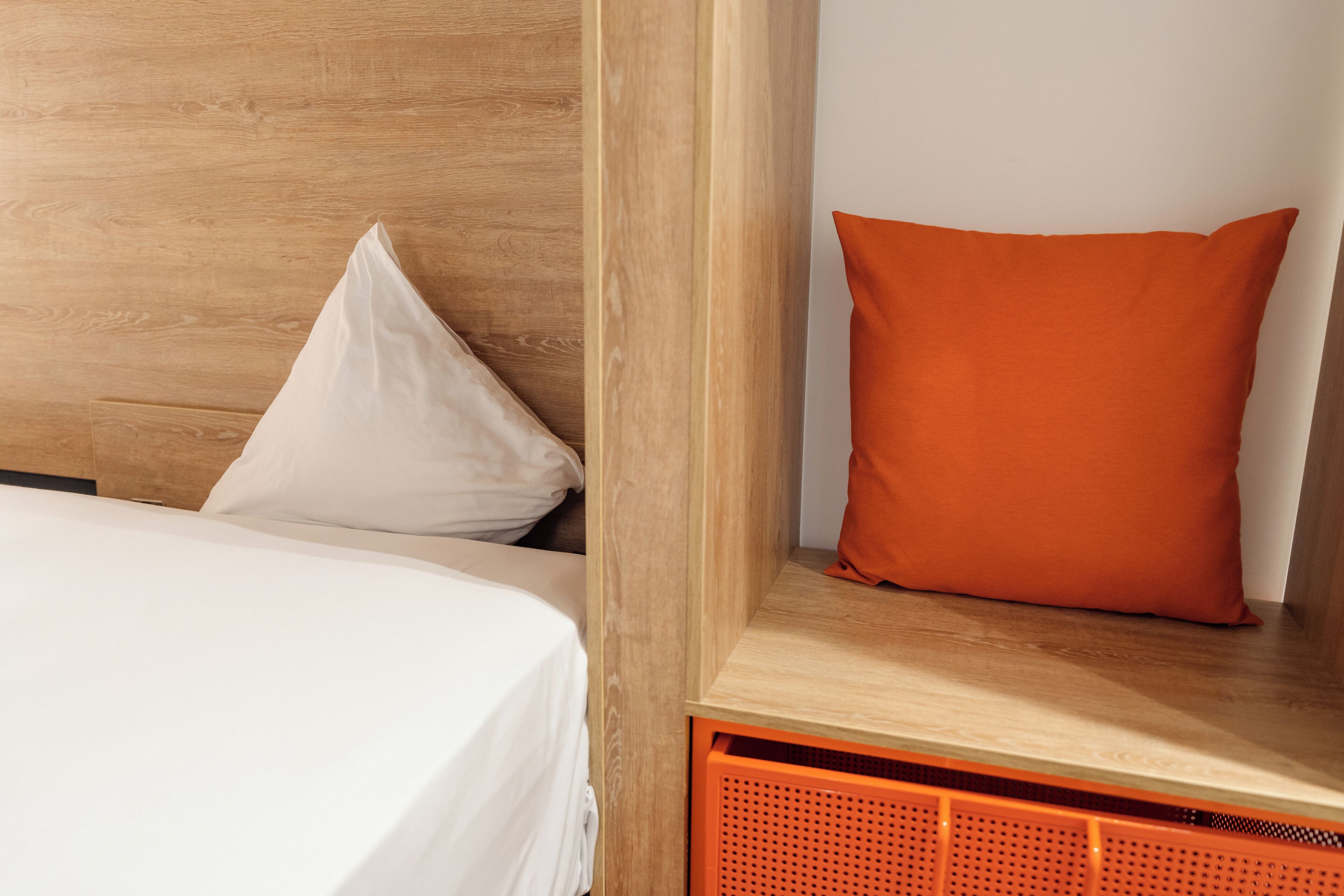 Un lit bien fait, avec une literie blanche, une tête de lit en bois et un coussin orange qui apporte une touche de couleur.