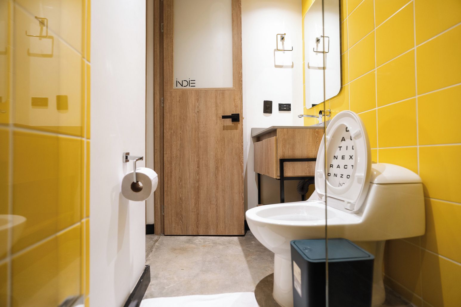 A bathroom with yellow tiles, a white toilet, a wooden door labeled 'INDIE', and a mirror.