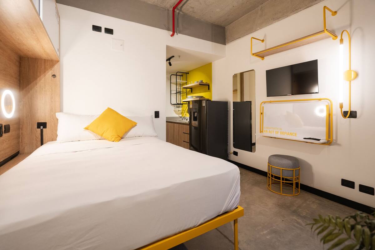 A modern hotel room with a bed, a TV, a small stool, and a kitchen area visible through an open door.