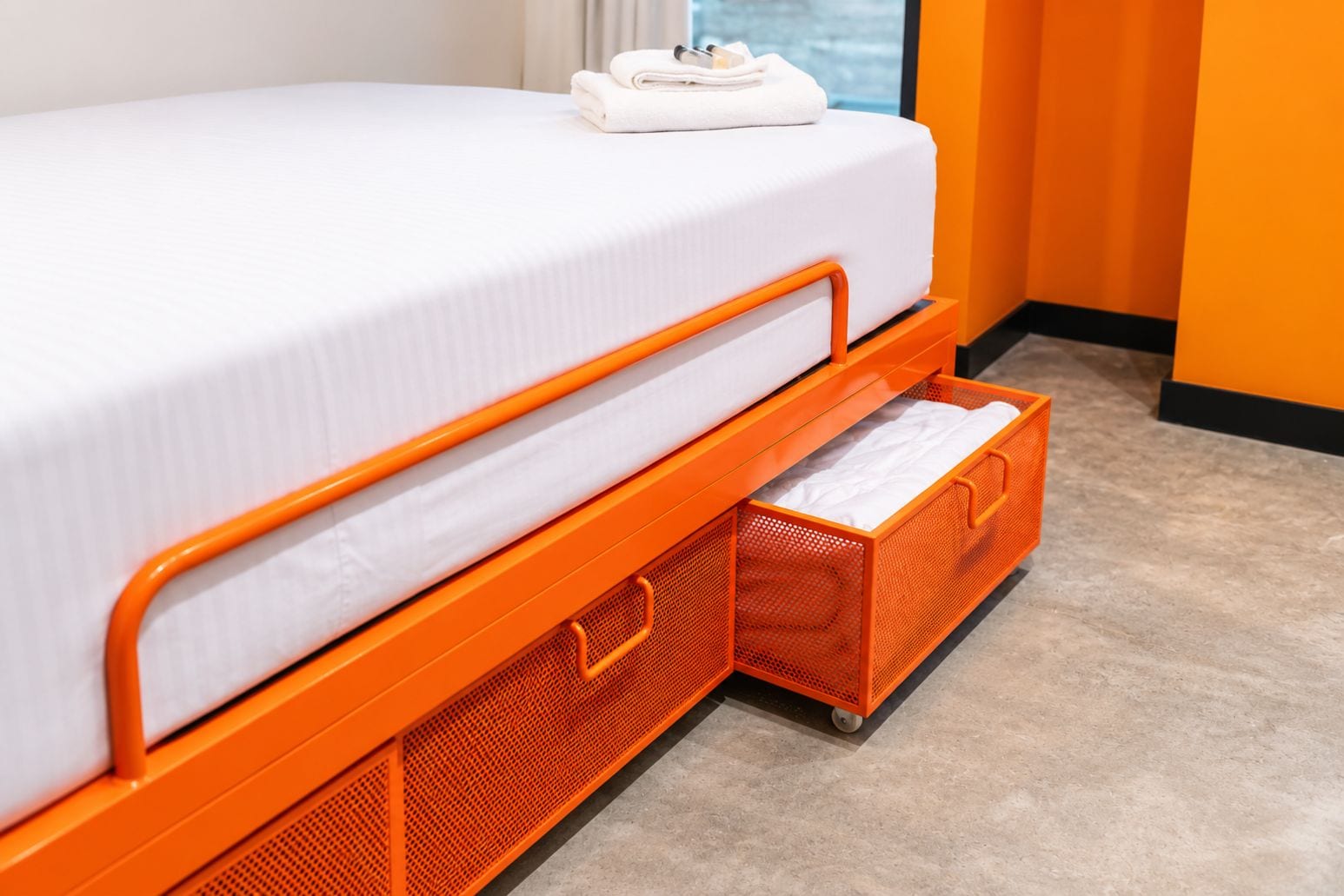 Una cama con estructura metálica naranja, ropa de cama blanca y dos cajones portaobjetos naranja debajo.