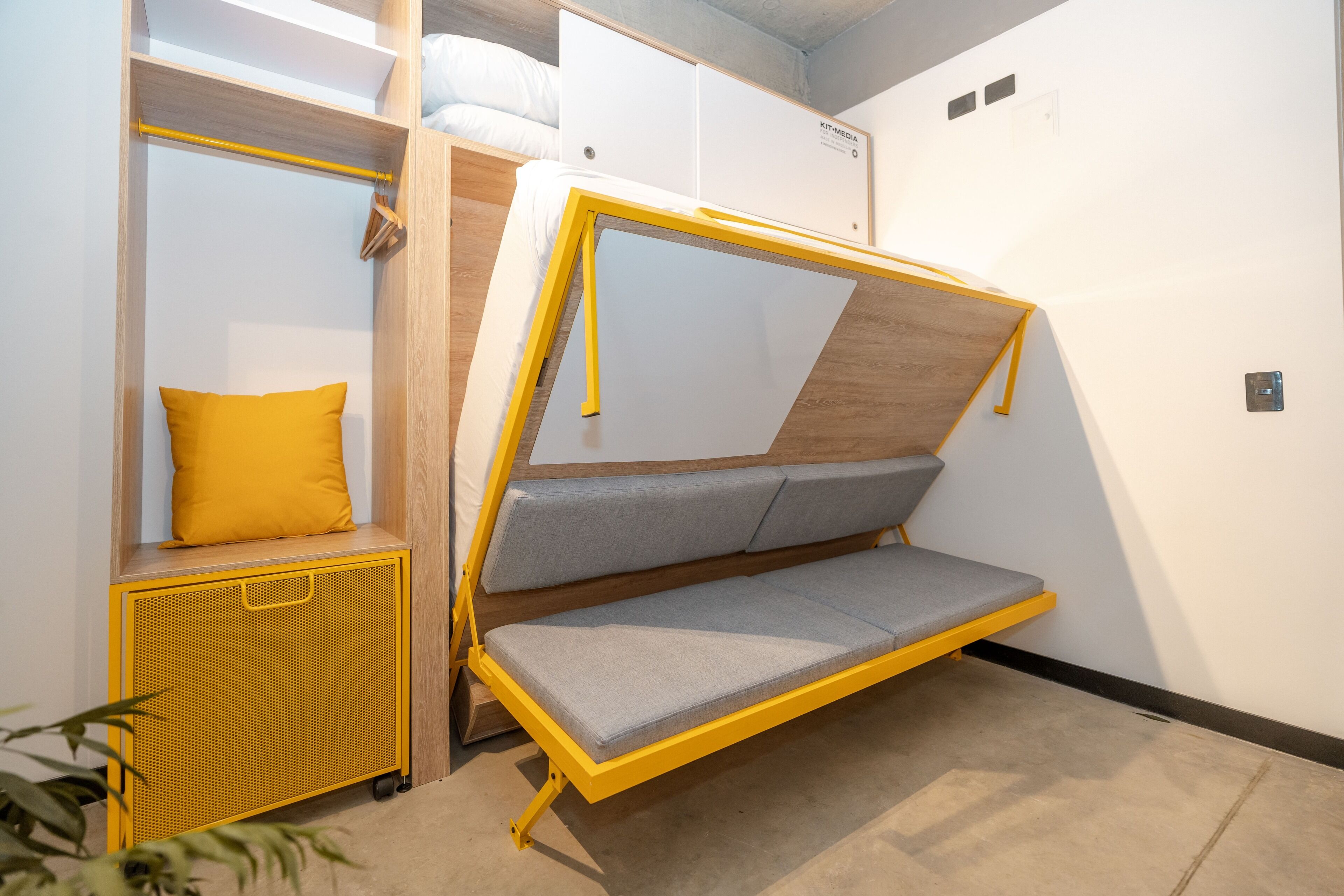 A compact room with a foldable bed, a small seating area with cushions, and a storage unit with a yellow mesh front.