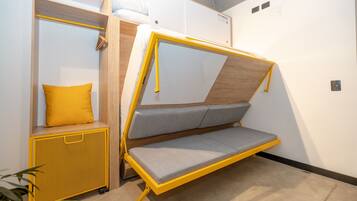 A compact room with a foldable bed, a small seating area with cushions, and a storage unit with a yellow mesh front.