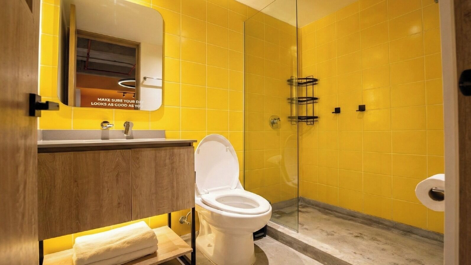 A bathroom with a yellow tiled wall, a white toilet, a glass shower enclosure, and a wooden vanity with a mirror.