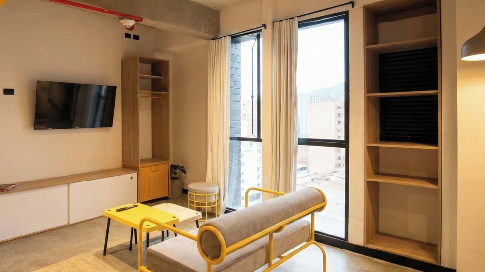 Una habitación moderna con televisión, una mesa amarilla, un banco y un amplio ventanal con cortinas.