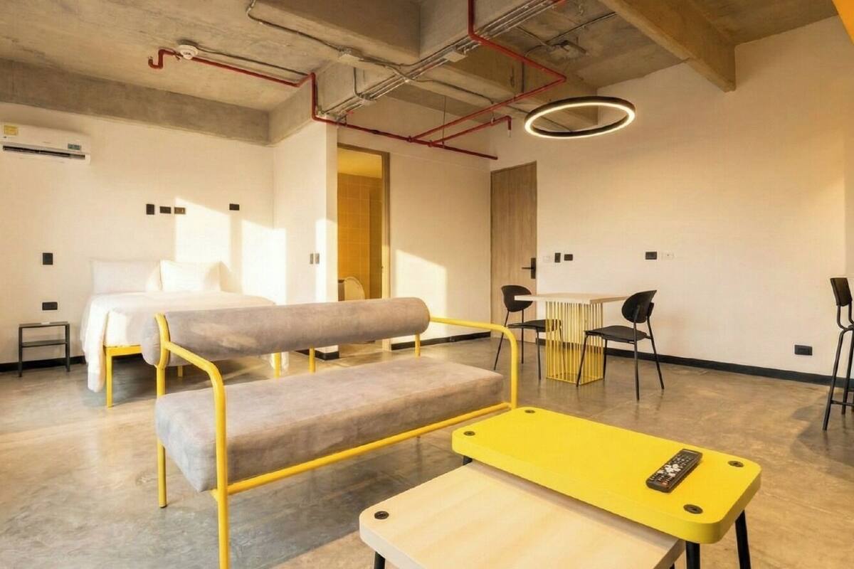 A modern hotel room with a bed, a bench, a small table, and a desk. There is a window, a door, and a ceiling with exposed pipes.