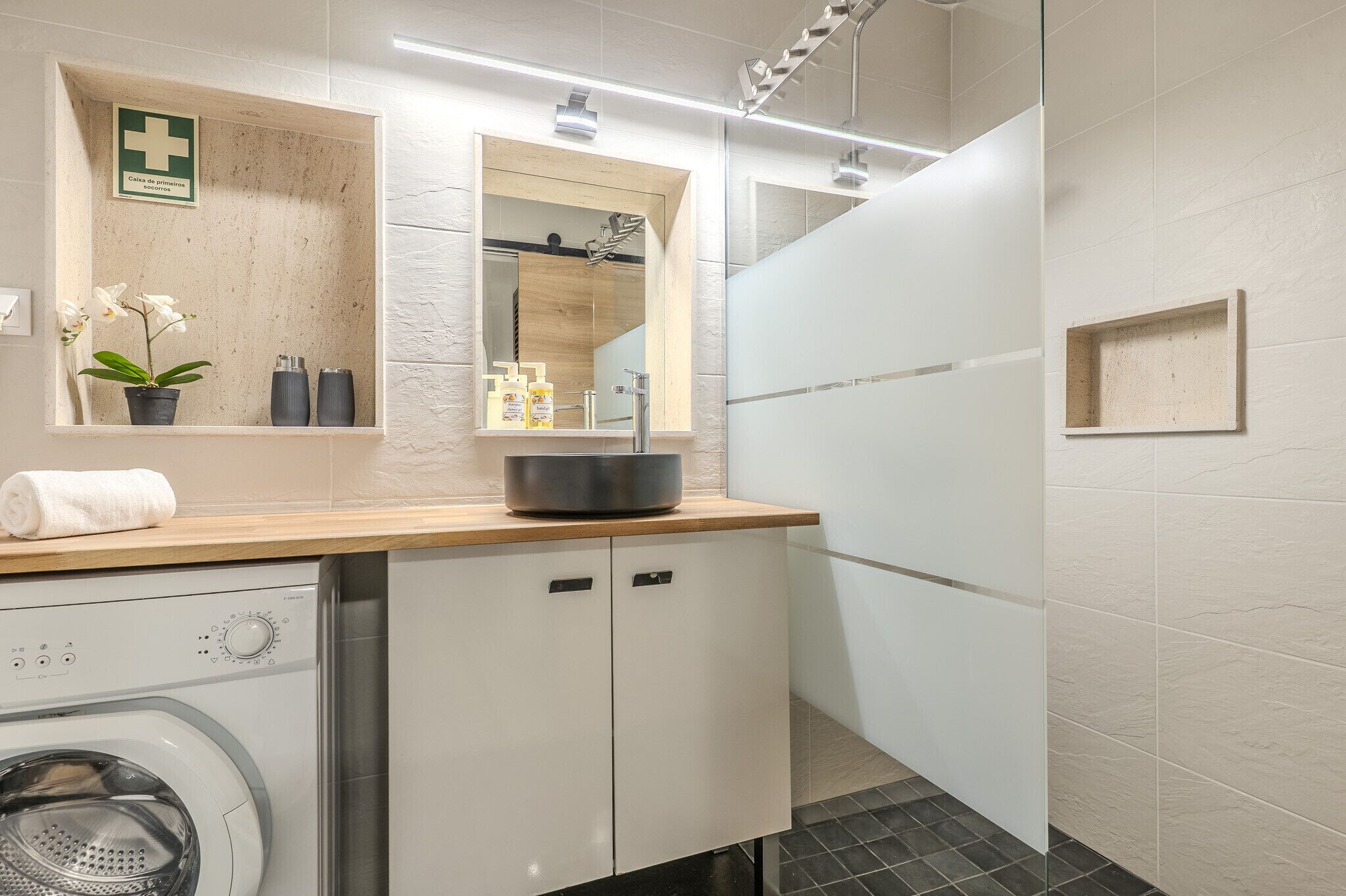 Een moderne badkamer met een wasmachine, een wastafel met spiegel en een klein plankje met toiletartikelen.