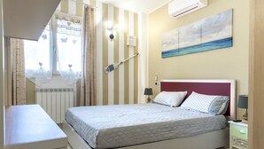 A bedroom with a bed, bedside tables, a window with curtains, a wall-mounted air conditioner, and a painting of a seascape. - N213 - Numana, new apartment in the center (Numana)