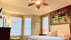 A bedroom with a bed, a nightstand, a lamp, a ceiling fan, and a tapestry on the wall.