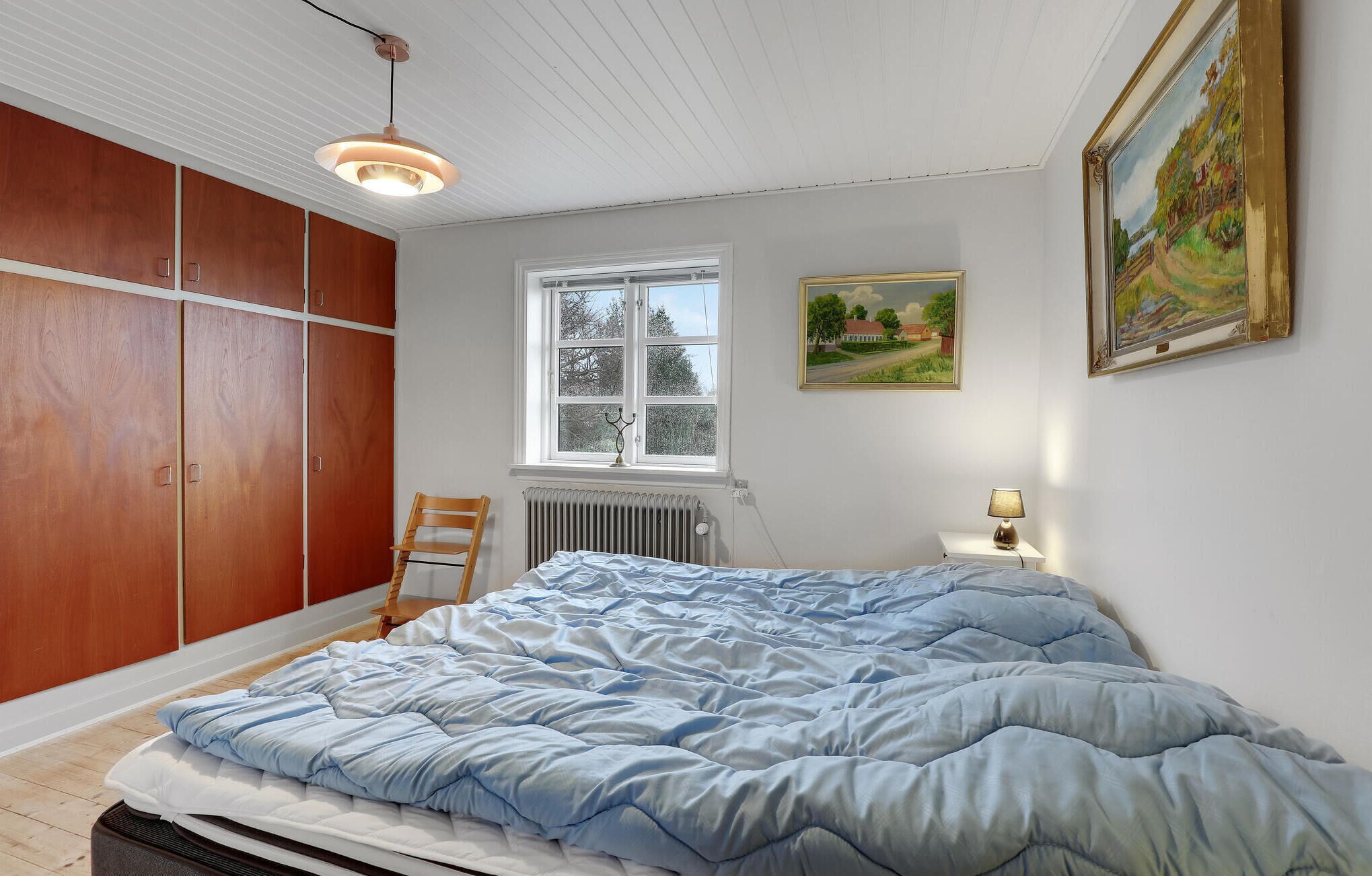 A bedroom with a bed, wooden wardrobe, window, radiator, and a painting on the wall.