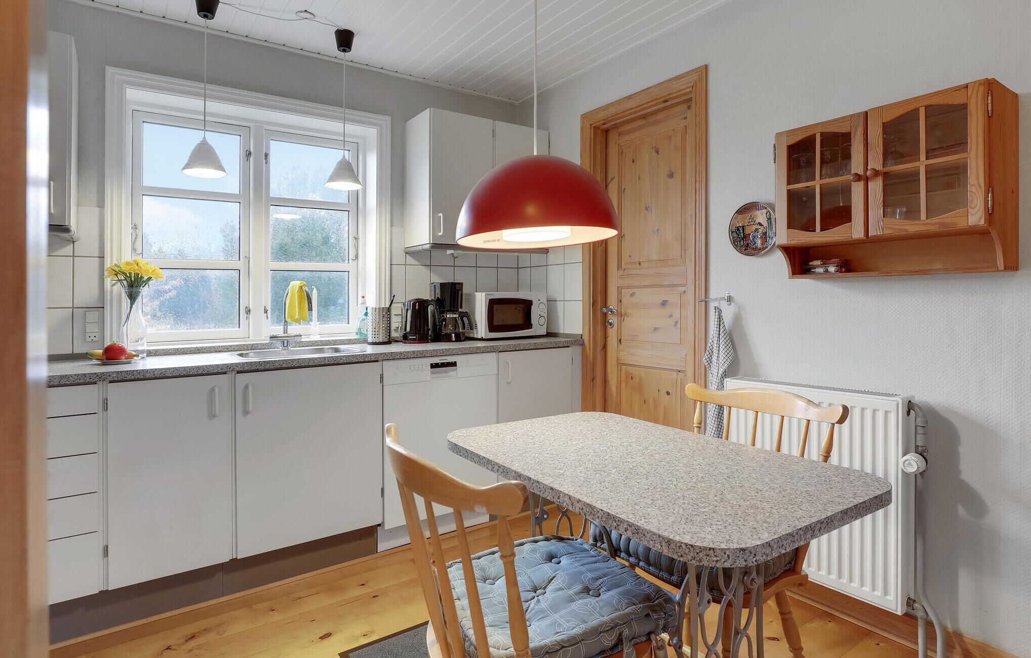 Une cuisine avec des meubles blancs, un plan de travail en granit, un luminaire suspendu rouge, une porte en bois et un petit meuble avec des portes en verre.