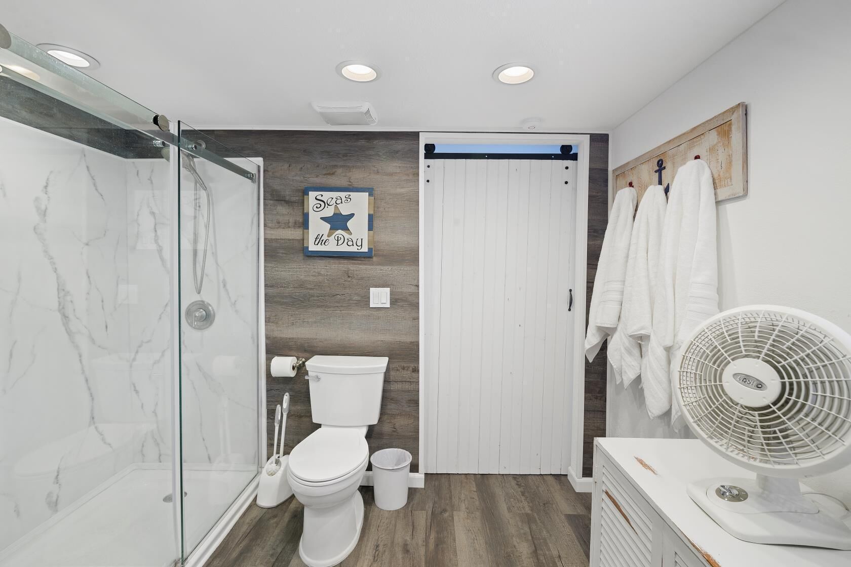 Ein Badezimmer mit Duschabtrennung aus Glas, Toilette, Wandventilator und Handtuchhalter mit Handtüchern.