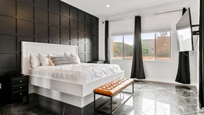 A modern bedroom with a large bed, a bench, a nightstand, a ceiling fan, and a window with curtains. - Baxter: Pet Friendly + Private Pool/Hot Tub + Golf (Hurricane)