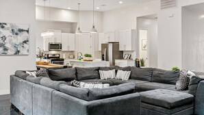 A modern living room with a grey sectional sofa, a dining area, and a kitchen with white cabinets and stainless steel appliances. - Baxter: Pet Friendly + Private Pool/Hot Tub + Golf (Hurricane)