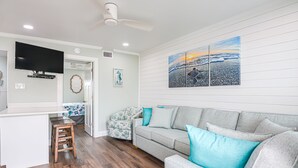 Uma sala de estar com um sofá cinzento, um banquinho de madeira, uma TV de tela plana e um quadro com uma cena de praia.