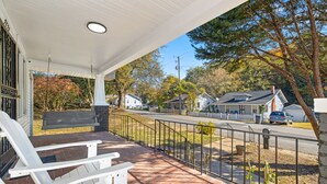 A covered porch with two white Adirondack chairs, a brick patio, and a view of a residential street with houses and trees.