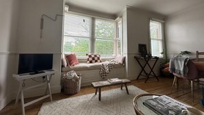 A cozy living room with a television, a bench with cushions, a small table, and a window with blinds. - Stylish Apt London Excel Olympic Close to Station (London)