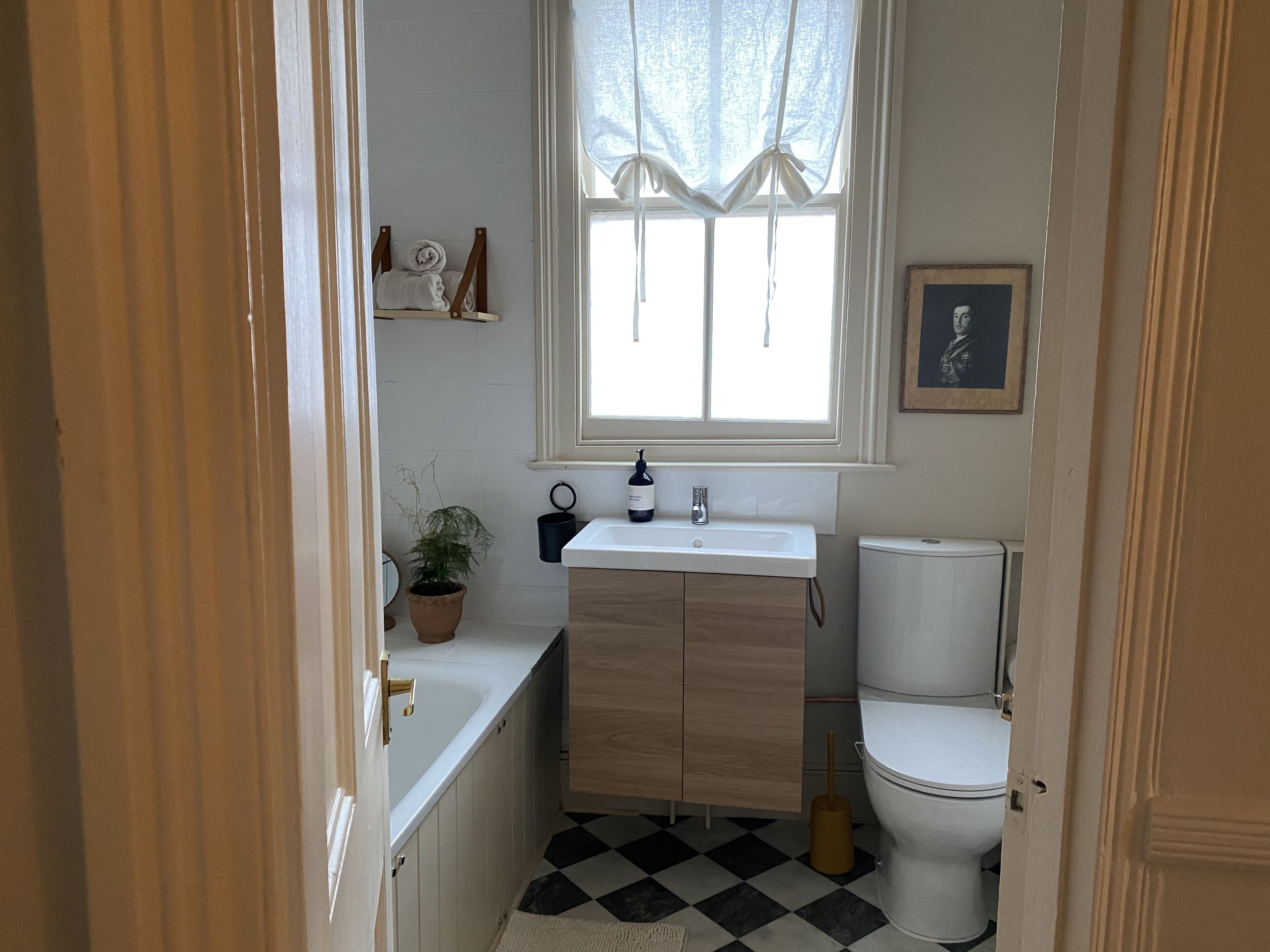 A small bathroom with a white sink, wooden cabinets, a toilet, a potted plant, and a window with sheer curtains.