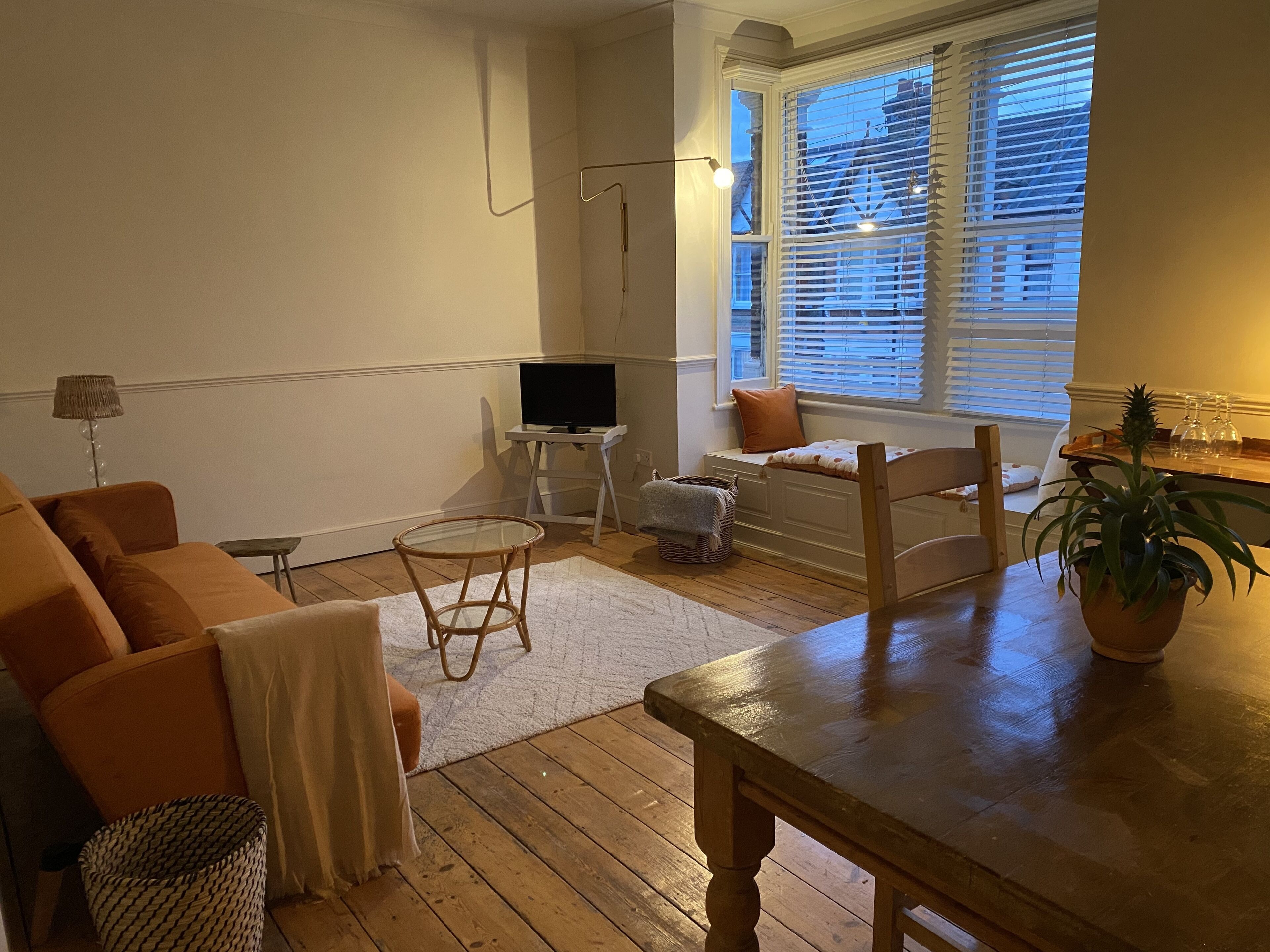 A living room with a wooden table, a sofa, a TV on a stand, a window with blinds, and a potted plant.