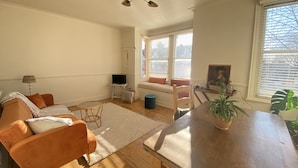 A living room with a sofa, a coffee table, a TV on a stand, and a window with blinds. - Stylish Apt London Excel Olympic Close to Station (London)