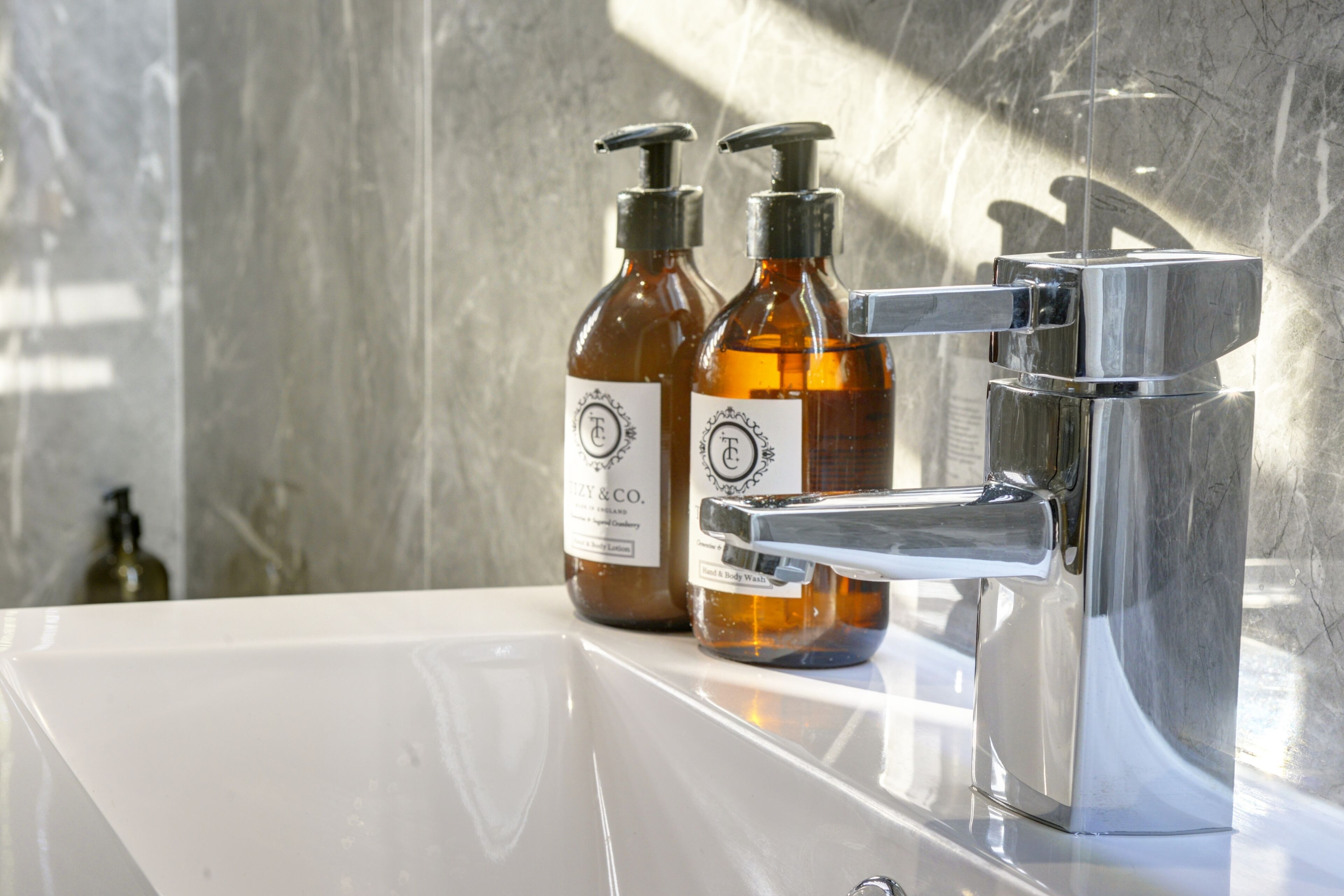 Un baño con lavamanos blanco, grifo cromado y dos botellas ámbar de jabón de baño sobre la encimera.