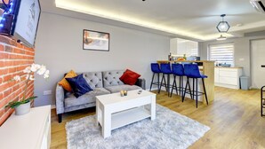 A modern living room with a grey sofa, white coffee table, and a kitchen area with blue bar stools. - Modern 2BR House & Private Parking & Kid-friendly (Rugby)
