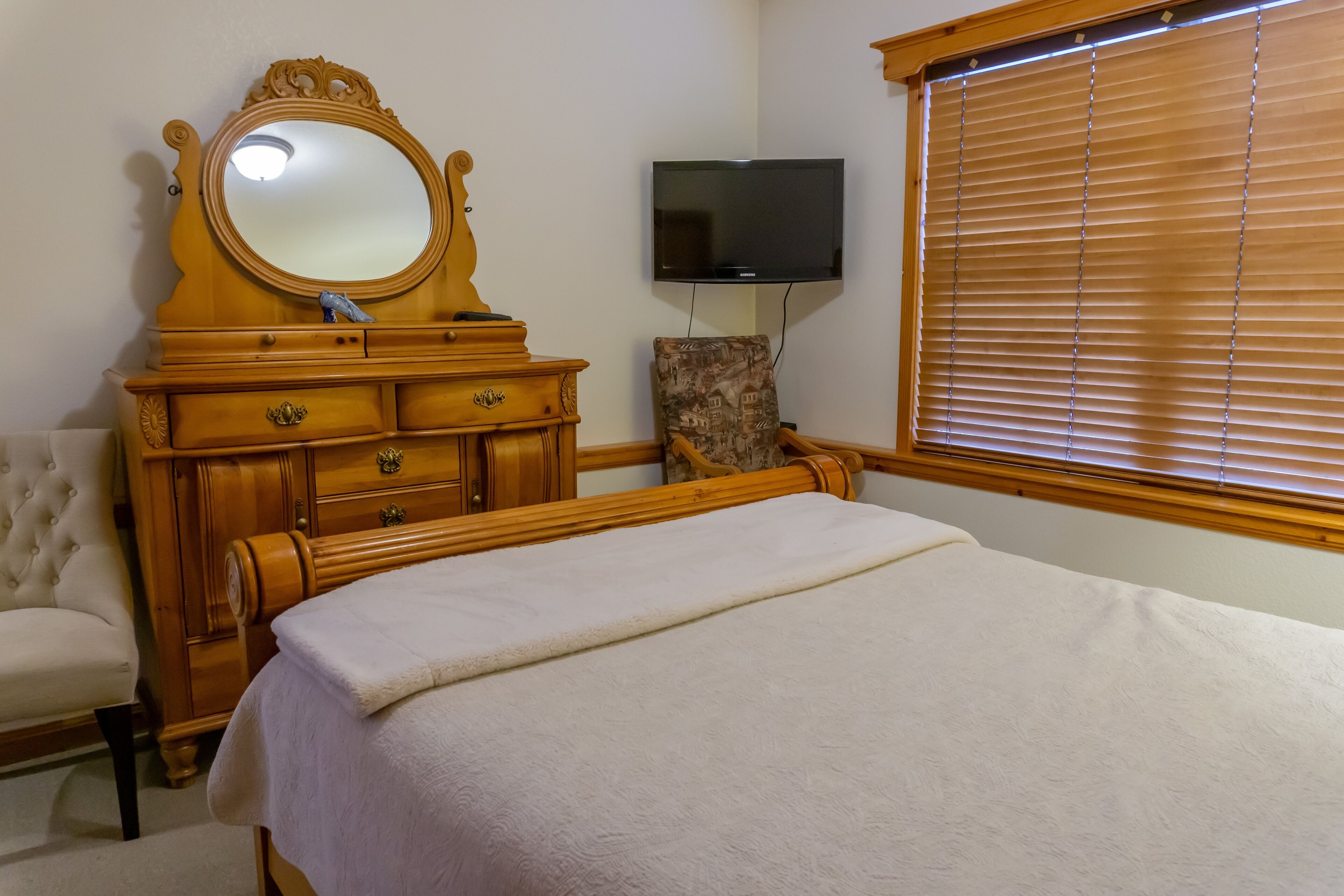 A bedroom with a wooden bed, a dresser with a mirror, a television, and a window with blinds.