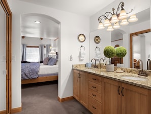 A bedroom with a bed, a nightstand, a dresser, and a bathroom with a sink, mirror, and lights.
