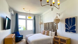 Una habitación de hotel moderna con una cama grande, televisión, escritorio, un sillón azul y una ventana con cortinas.