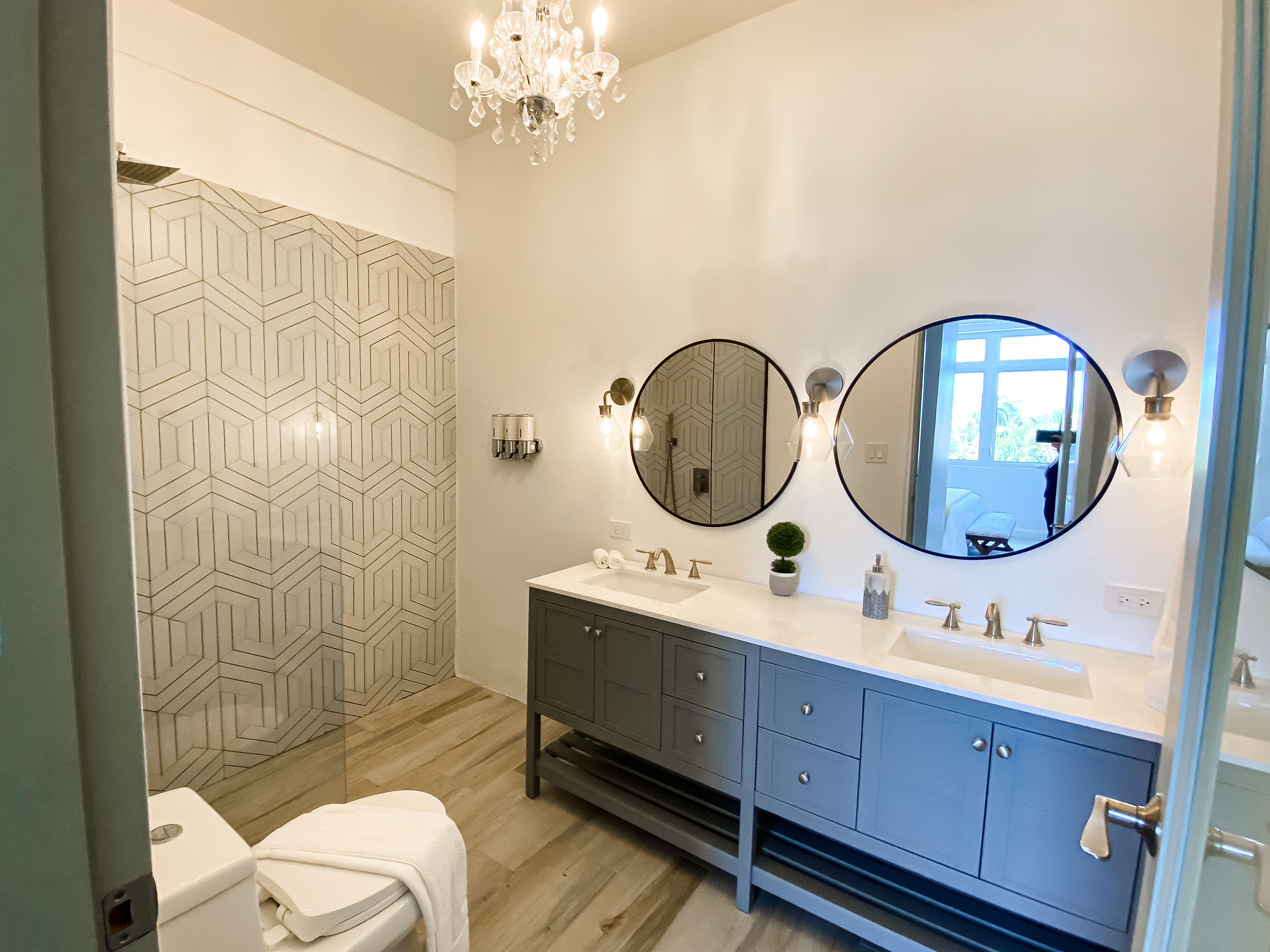 Un baño moderno con tocador doble, dos espejos grandes, una araña y pared de ducha con diseño geométrico.