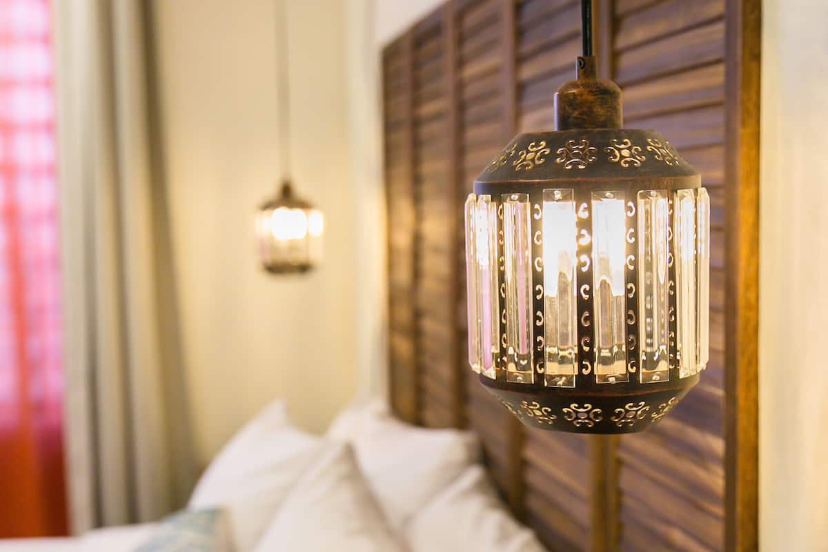 Uma cama com lençóis brancos e cabeceira de madeira, luminária pendente com padrão decorativo e uma janela com cortinas.