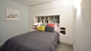 A bedroom with a bed, built-in storage, and a wall-mounted shelf with books and decorative items.