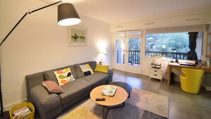 A living room with a sofa, coffee table, and a desk with a chair. There is a window with a view of trees and a balcony.
