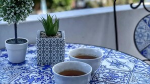 Una pequeña mesa con dos tazas blancas de té sobre baldosas azules y blancas, una planta en maceta y una silla de metal decorativa.