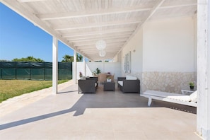 A covered outdoor patio with a stone wall, a white bench, and a dining area with chairs and a table.
