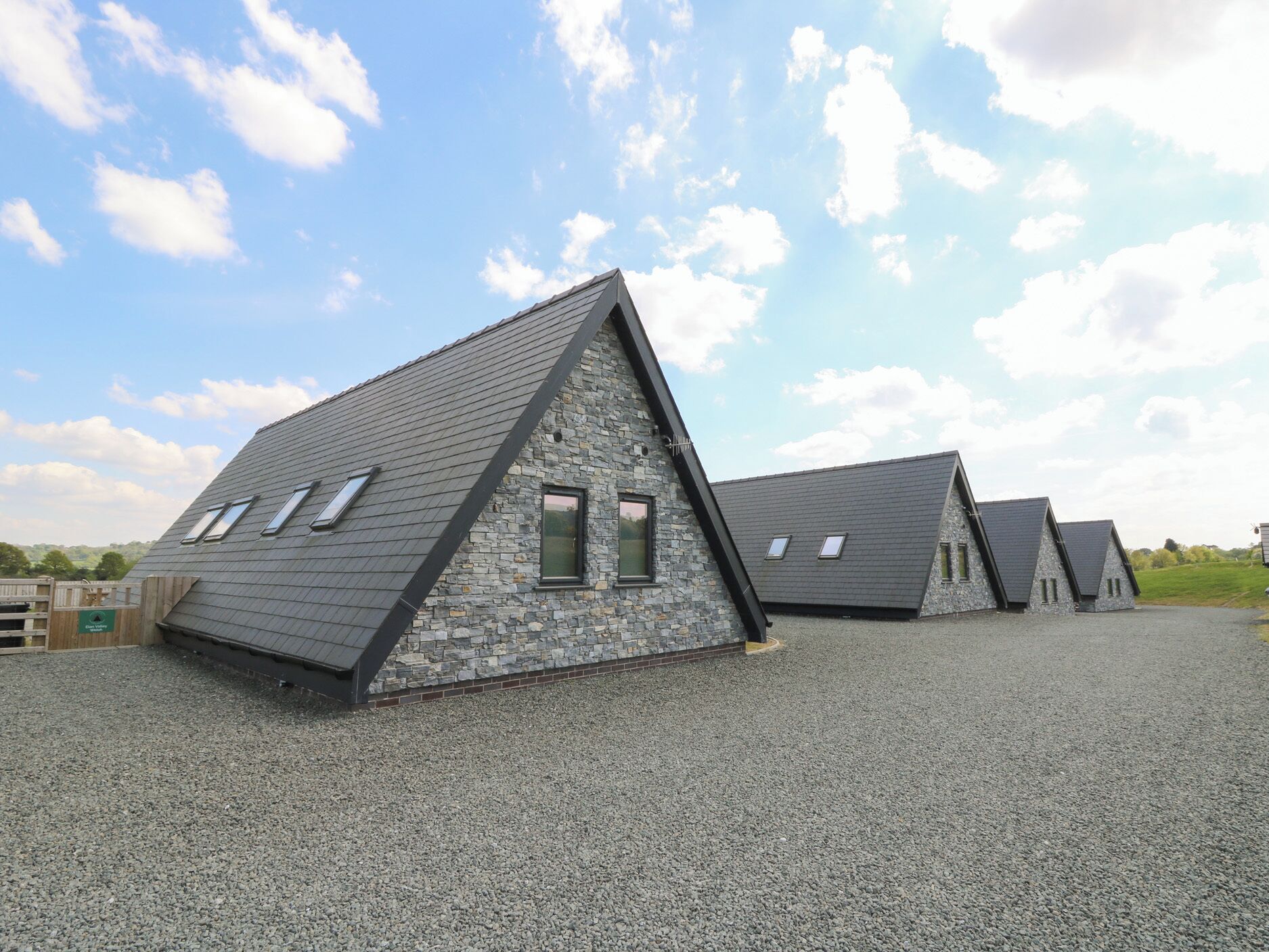 A row of modern, triangular-roofed buildings with stone facades and multiple windows, set on a gravel surface under a clear sky.