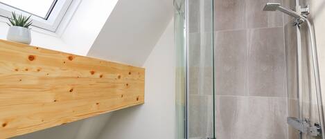 Una ducha moderna con puerta de vidrio, un estante de madera con una planta y un tragaluz arriba.