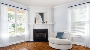 Una sala de estar con chimenea, un sillón blanco y ventanas grandes con persianas.