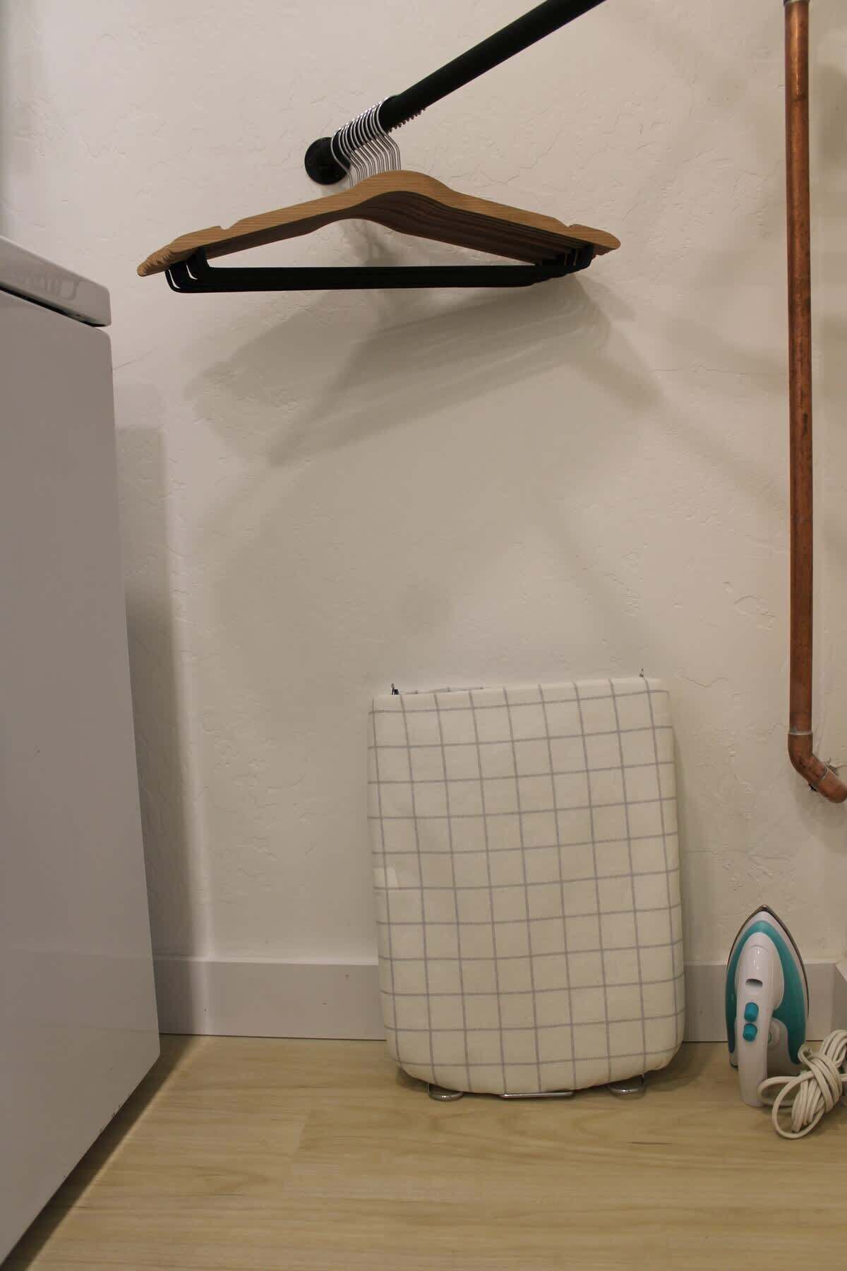 Un coin de pièce avec un radiateur blanc, un portant à vêtements suspendu, une planche à repasser blanche et un tuyau en cuivre.