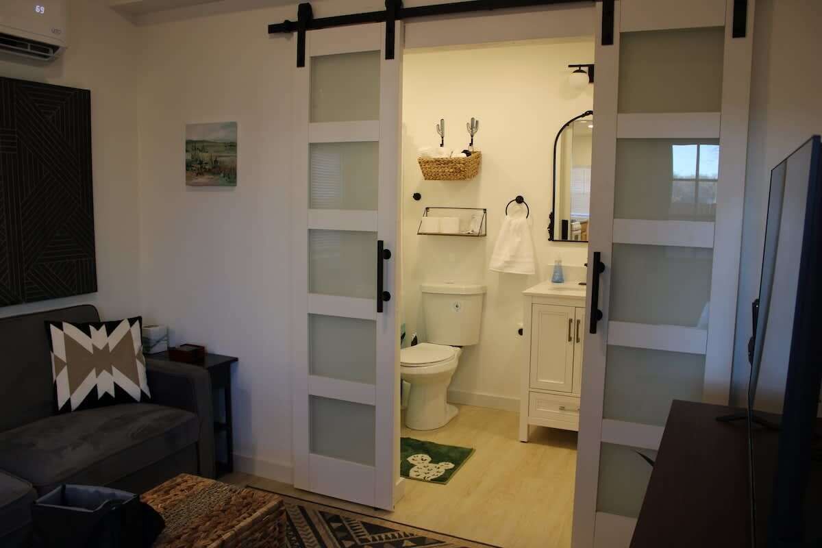 En stue med sofa, sofabord og tv. En skydedør fører til et badeværelse med toilet, vask og spejl.