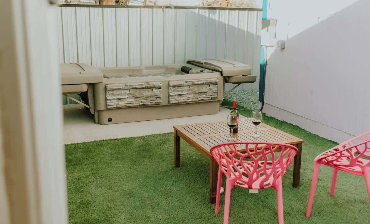 A small outdoor patio with a wooden table and two pink chairs, a beige sofa, and a hot tub.