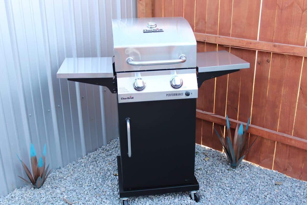 A Char-Briill portable grill with a performance feature, placed on a wheeled base, against a wooden fence and corrugated metal wall.