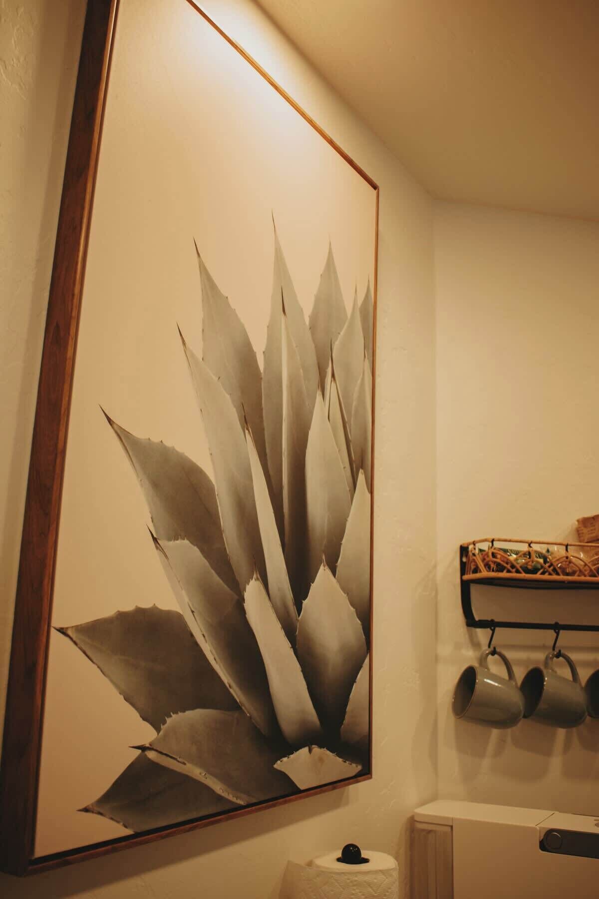 A bathroom with a large wall-mounted artwork of agave plants, a toilet, and a shelf with mugs.