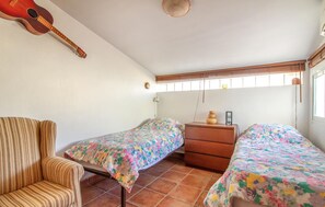 A room with two beds, a chair, a dresser, a guitar, and a window with blinds.