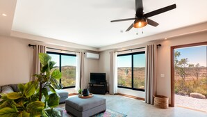 A spacious living room with a grey sofa, a small ottoman, a ceiling fan, and a large window offering a view of the desert landscape.