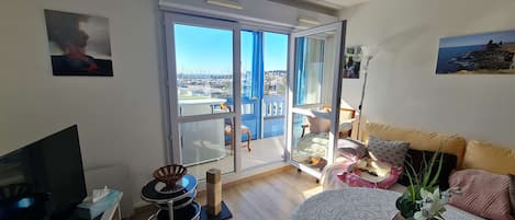A modern hotel room with a large sliding glass door leading to a balcony, a flat-screen TV, a small round table, and a bed with a patterned bedspread and pillows.