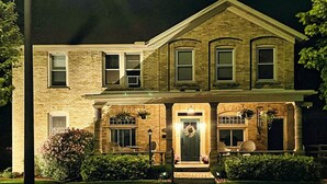 A two-story brick house with a front porch, a central door with a wreath, and a small garden area. - Home Port Vacation Escape, Luxury Historic Home, Walk to Port, Beach, Shops (Port Washington)