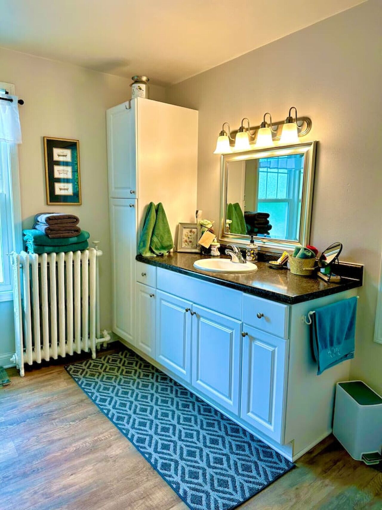 A bathroom with a white radiator, a sink with a mirror above it, and a blue patterned rug on the floor.