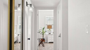 A hallway with a wooden floor, white walls, and a door leading to a room with a bed and a plant.