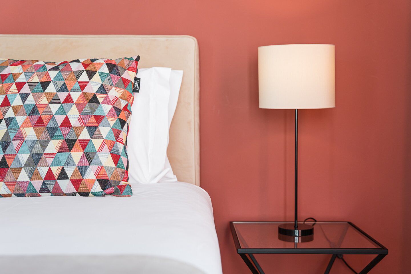 A neatly made bed with a colorful geometric-patterned pillow and a white pillow, a bedside table with a lamp, and a plain red wall.